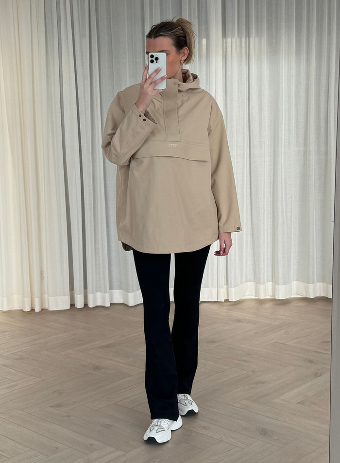A woman wearing a Beige Waterproof Oversized Anorak from aim'n, paired with black leggings and white sneakers while taking a mirror selfie.