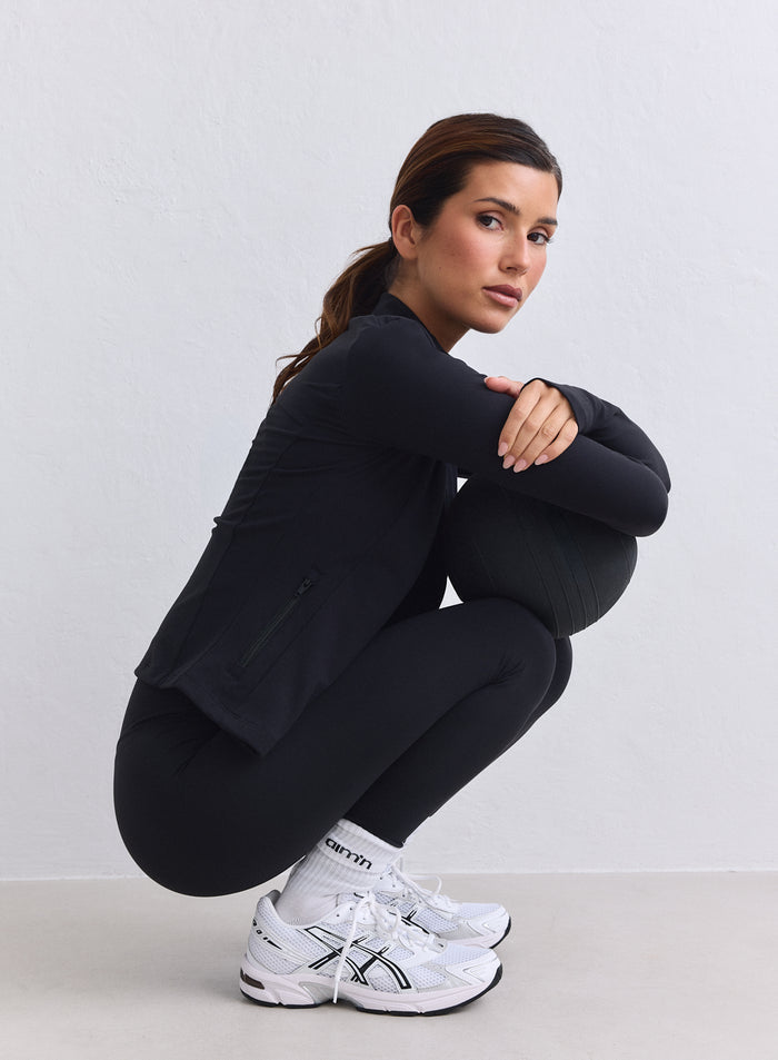 Woman in a black aim'n Sense Zip Jacket and leggings with a second skin feel, crouching with a ball. [XS-165cm]