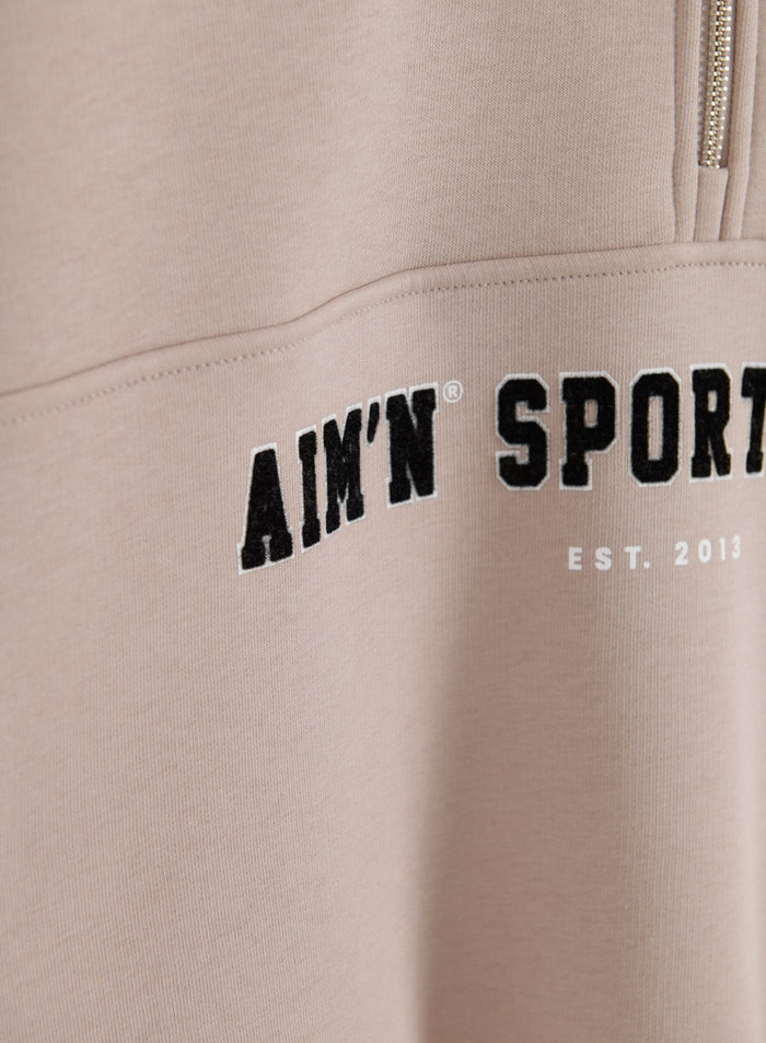 Varsity-style aim'n embroidery on a dark clay sweatshirt with a metallic half-zip detail.