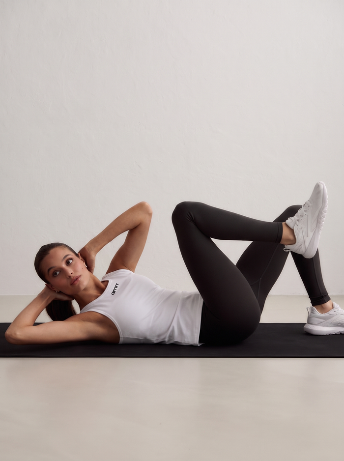 A model lying on a mat performing core exercises, wearing a white Soft Basic Singlet, black leggings and white sneakers [S-174 cm]